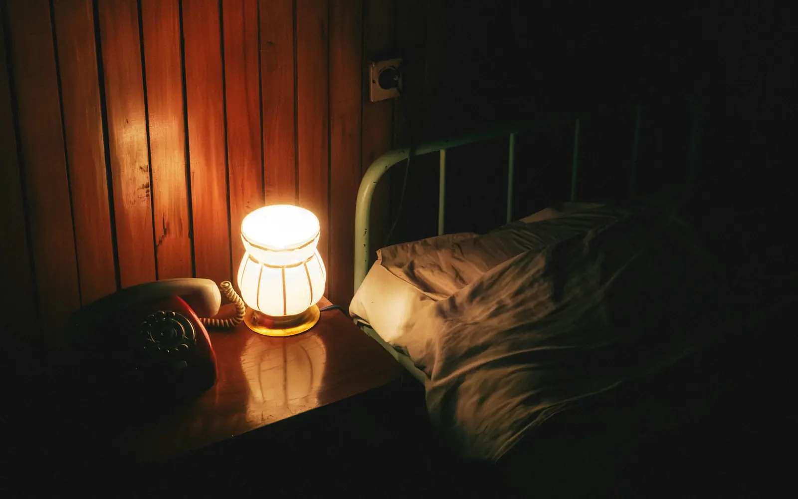 A small lamp glowing beside a bed and an old telephone at night